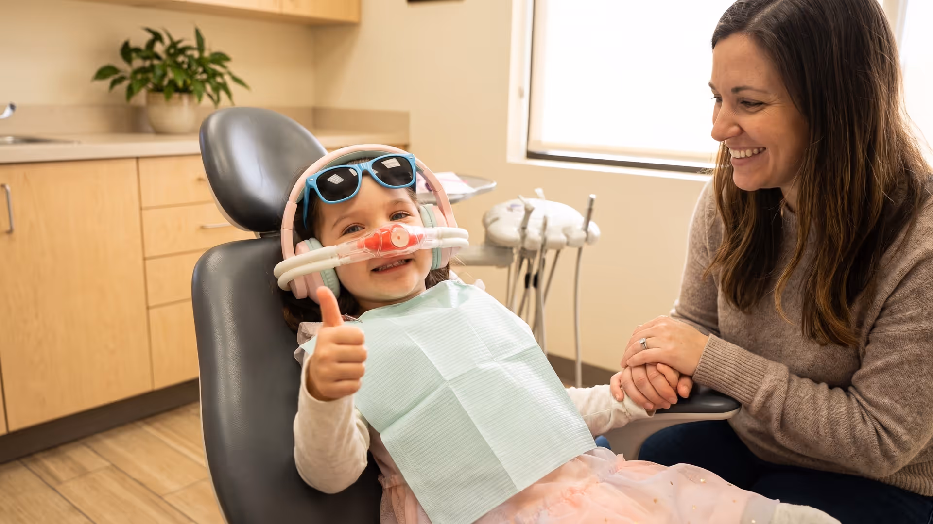 A smiling girl giving a thumbs-up in the dental chair while relaxed on nitrous oxide, her mother holding her hand